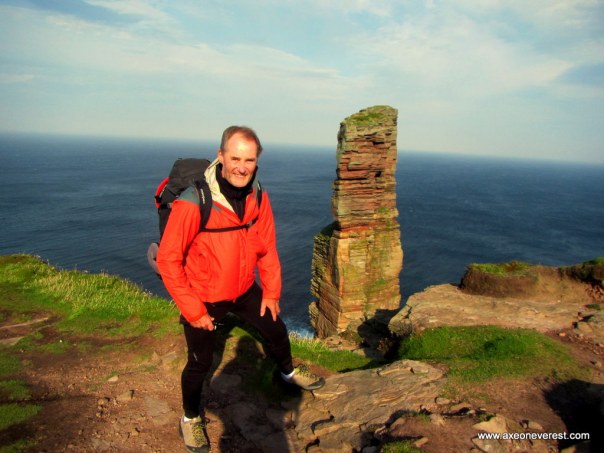 Alan Silva standing on the sea cliffs in strong winds - the summit of the Old Man of Hoy seen behind him.